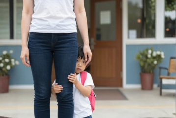 Mädchen versteckt sich hinter den Beinen ihrer Mutter. Sie sieht schüchtern aus und trägt einen Rucksack. Sie stehen vor dem Eingang der KiTa