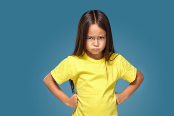 Kind stützt die Arme in die Hüften und guckt böse. Blauer Hintergrund.