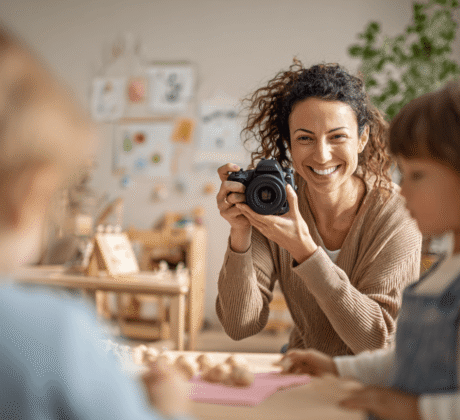Frau mit nach hinten zusammengesteckten Locken lacht Kinder freundlich an und macht ein Foto von ihnen.