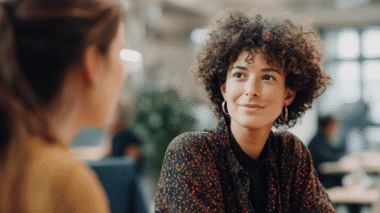 Junge Bewerberin mit dunklen Locken, einer gepunkteten Bluse und goldenen Ohrringen im Bewerbungsgepräch mit der Kita-Leitung