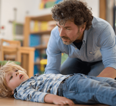 Kind liegt auf dem Boden in der Kita. Neben ihm kniet ein Erzieher. Symbol für Ersthelfer in der Kita