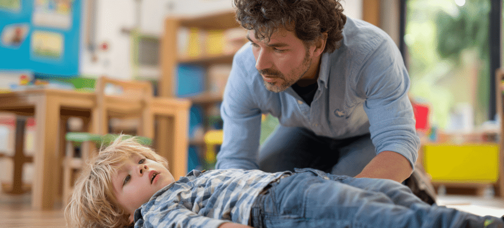 Kind liegt auf dem Boden in der Kita. Neben ihm kniet ein Erzieher. Symbol für Ersthelfer in der Kita