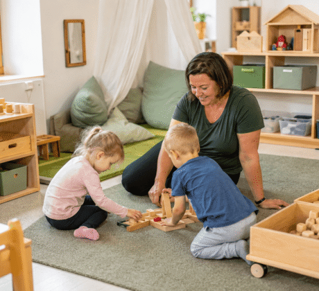 Erzieherin spielt mit zwei Kindern auf einem Spielteppich. Der Raum ist bedürfnisorientiert eingerichtet mit viel Holz und klarer Struktur