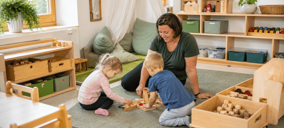 Erzieherin spielt mit zwei Kindern auf einem Spielteppich. Der Raum ist bedürfnisorientiert eingerichtet mit viel Holz und klarer Struktur