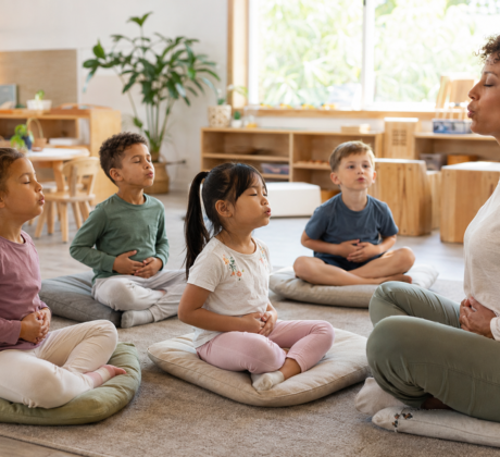 Kinder auf Sitzkissen im Kita-Gruppenraum machen Yoga-Atemübung mit Erzieherin
